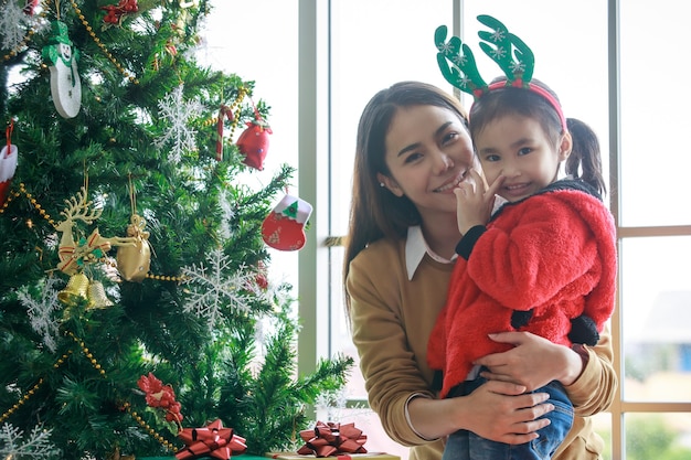 Feliz madre de familia asiática abraza y enseña a su hija en suéter rojo y diadema de cuerno de reno que decora el árbol de pino de Navidad con artículos colgantes de brillo que celebran el evento de víspera de Navidad en la sala de estar de casa.