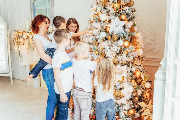 Feliz madre de familia y cinco hijos decorando el árbol de Navidad en la víspera de Navidad en casa. Hijos de hijas de mamá en habitación luminosa con decoración de invierno. Navidad año nuevo tiempo de celebración.