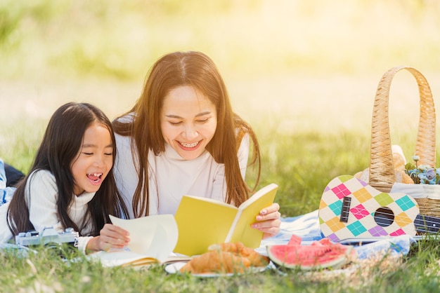 Feliz madre de familia joven asiática y niña pequeña divirtiéndose y disfrutando al aire libre en una manta de picnic leyendo un libro en el parque de primavera del jardín de verano, concepto de relajación familiar