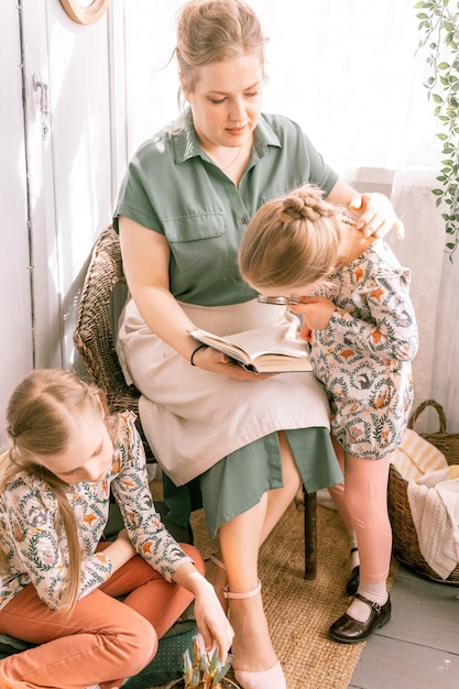 Feliz madre de familia con niños pequeños, hijas, niñas, se divierten juntas en la casa de campo de los suburbios en un día soleado de verano, mamá sincera abraza y lee un libro a un niño explorando un libro con una lupa