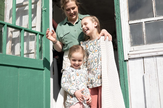 Feliz madre de familia con niños pequeños, hijas que ayudan a las niñas, se divierten en la vida rural de los suburbios el día de la lavandería. Mamá sincera abraza y cubre a los niños con cuadros y hace las tareas del hogar en el porche de la casa de campo.