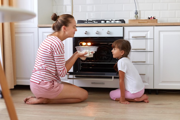 Feliz madre joven y linda hija horneando pastel o pastel en la cocina madre e hijo oliendo un delicioso postre sabroso gente sentada en el suelo disfrutando del sabor