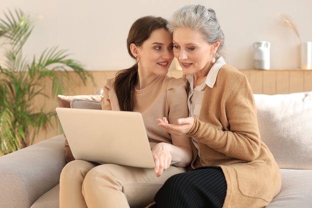 Feliz madre media anciana sentada en el sofá con su hija, mirando la computadora portátil. Mujer joven mostrando video, fotos a mami, relaciones de confianza. Concepto de familia.