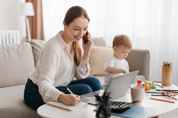 Feliz madre de negocios trabajando en casa mientras su pequeño hijo juega al lado de su señora hablando por teléfono