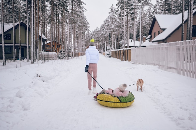 Feliz madre y niña linda en ropa rosa cálida caminando divirtiéndose paseos en tubo de nieve inflable con perro shiba inu rojo en campo de invierno blanco como la nieve al aire libre