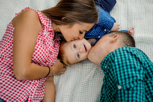 Feliz madre y padre besando a su hijo vestido con las camisas a cuadros que yacen en la tela escocesa en el parque