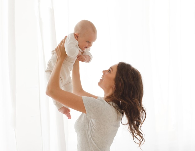 Feliz madre sonriente con bebé divirtiéndose juntos en el blanco de las cortinas del fondo de la ventana