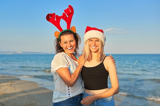 Feliz madre sonriente e hija adolescente con sombrero de Santa Claus en la playa