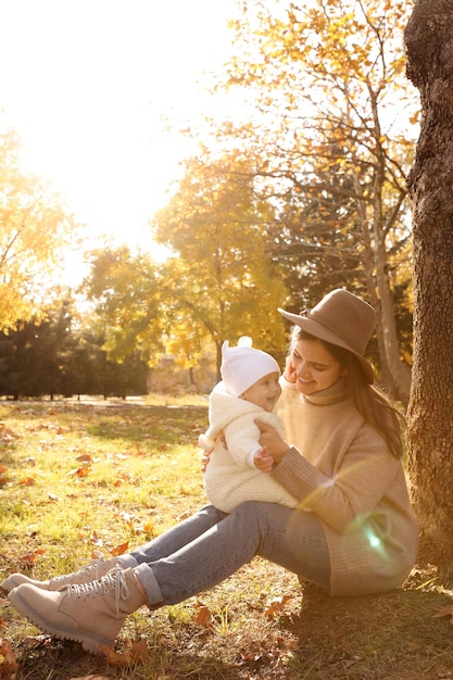 Feliz madre con su hija pequeña sentada cerca de un árbol en el parque en el soleado día de otoño espacio para texto