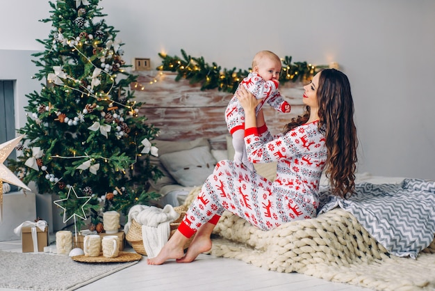 Feliz madre con su pequeña hija en ropa de vacaciones con ciervos impresos y copos de nieve divirtiéndose en la cama en una habitación acogedora