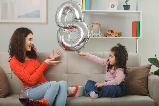 Feliz madre y su pequeña hija sentada en un sofá con un globo en forma de número ocho sonriendo alegremente divirtiéndose juntos en la sala de estar luminosa celebrando el día internacional de la mujer el 8 de marzo