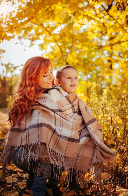 Feliz madre y su pequeño hijo caminando y divirtiéndose en el bosque de otoño. Mujer envuelve a su hijo en una bufanda