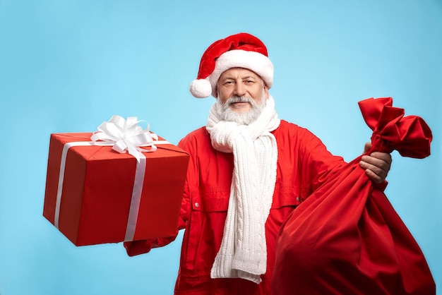 Feliz maduro barbudo papai noel com chapéu vermelho, jaqueta e lenço de malha branco segurando nas mãos a caixa de presente vermelha e um saco grande, isolado sobre fundo azul. presentes de feriado e época de natal.