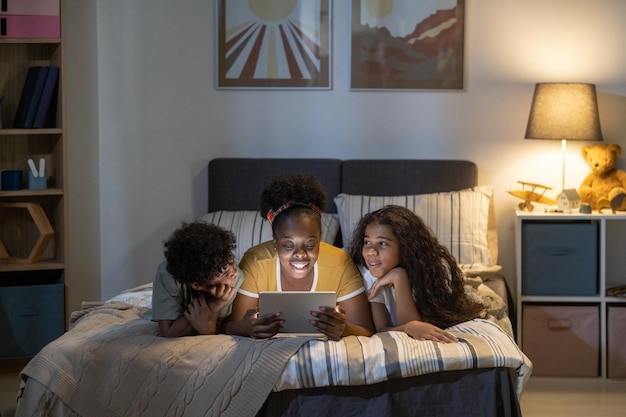 Feliz mãe afro-americana de cabelos cacheados deitada na cama e usando o tablet enquanto lê um conto de fadas