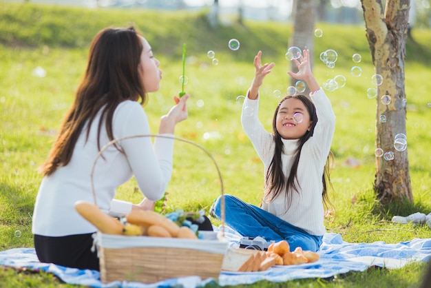 Feliz mãe asiática e filha pequena se divertindo e curtindo ao ar livre juntos sentados na grama soprando bolhas de sabão durante um piquenique no parque jardim em um dia ensolarado tempo para a família