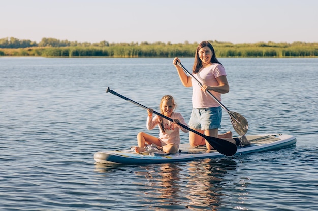 Feliz mãe caucasiana e filha embarcando com remos nas mãos olhando para a câmera e sorrindo no lago azul cristal com juncos verdes no fundo Estilo de vida ativo