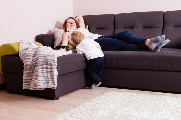 Feliz mãe deitado no sofá com seu filho bebê em pé perto dela. Jovem mãe deitada no sofá amamentando seu filho, conceito