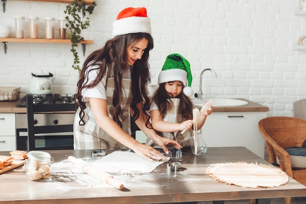 Feliz mãe e criança alegres com chapéus de Papai Noel estão cozinhando biscoitos de Natal na cozinha ano novo e Natal