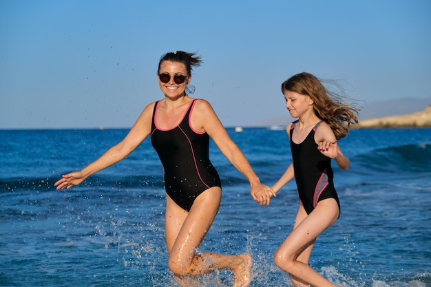 Feliz mãe e filha de meia idade correndo ao longo da praia, férias ativas no mar de verão juntos, fundo da paisagem do pôr do sol do mar. Família, alegria, esporte, estilo de vida, conceito de fim de semana