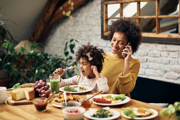 Feliz mãe negra falando ao telefone enquanto toma café da manhã com a filha na sala de jantar