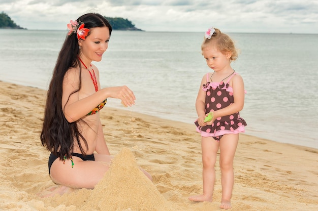 Feliz mãe sorridente com uma filha brinca na praia e constrói castelos de areia em um dia ensolarado de verão Mãe e menina criança Phuket Island Tailândia Karon Beach