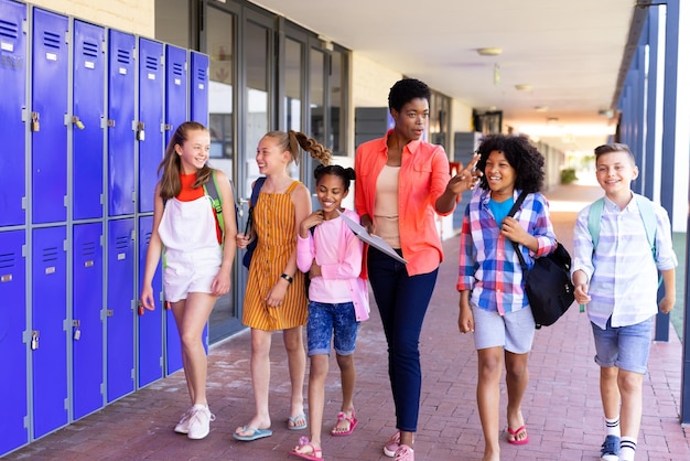 Foto feliz maestra diversa y niños de la escuela caminando juntos en el pasillo de la escuela y hablando