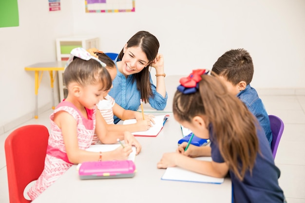 Feliz maestra de jardín de infantes ayudando a sus estudiantes con algunos ejercicios de escritura en el aula