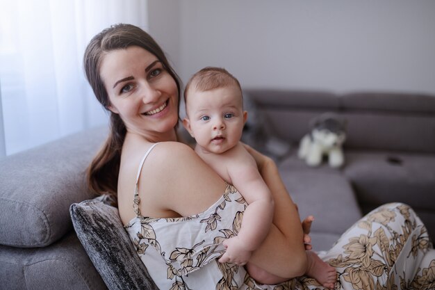 Feliz mamá caucásica sosteniendo a su adorable hijo mientras está sentado en el sofá. Bebé mirando con una pequeña sonrisa.