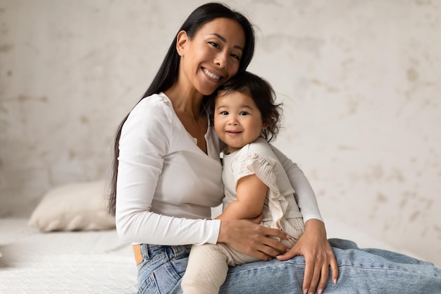 Feliz mamá coreana abrazando a una adorable hija pequeña sentada en casa