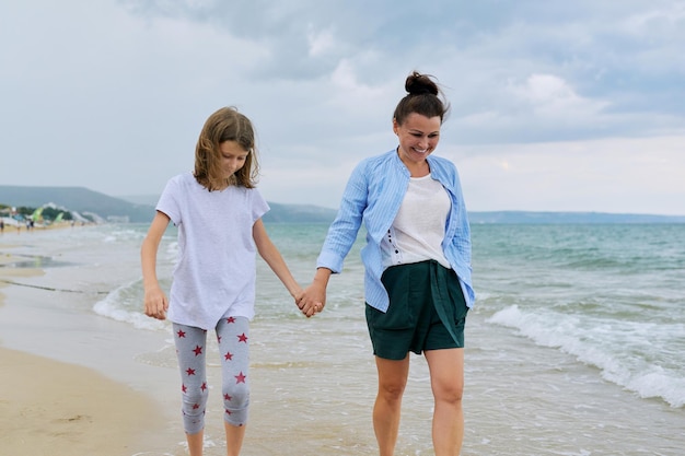 Feliz mamá e hija caminando en la playa del mar cogidos de la mano