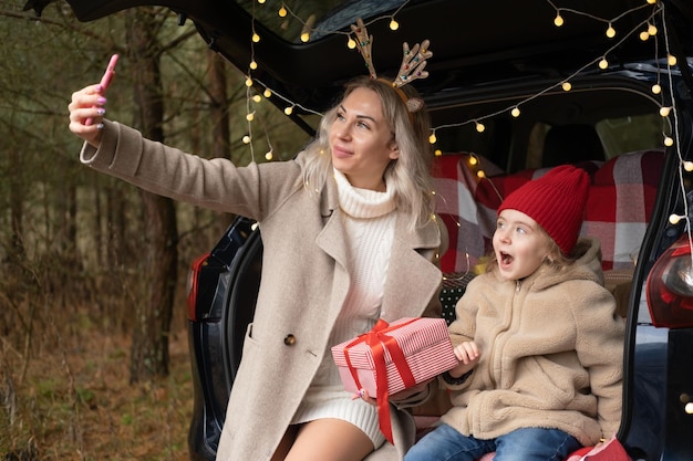 Feliz mamá e hija haciendo videollamadas con cajas de regalo de Navidad en un auto decorado con Navidad