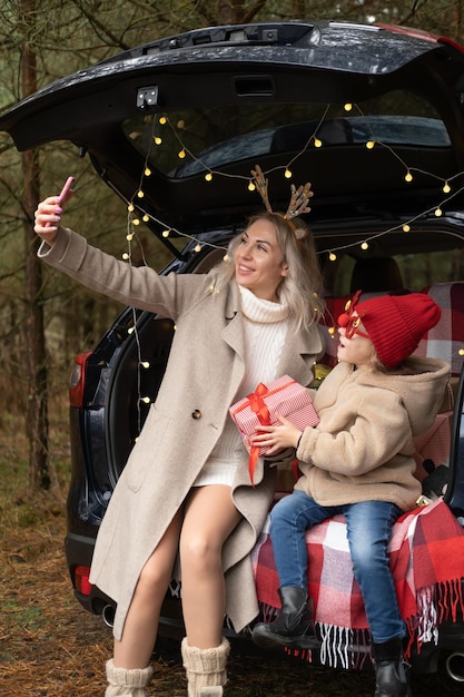 Feliz mamá e hija haciendo videollamadas con cajas de regalo de Navidad en un auto decorado con Navidad