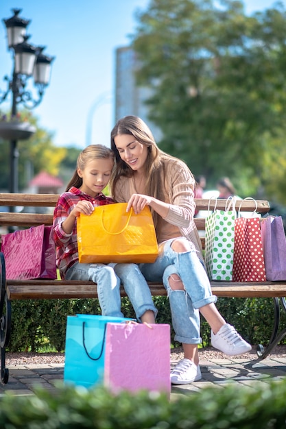 Feliz mamá e hija mirando dentro de la bolsa de compras en un banco del parque