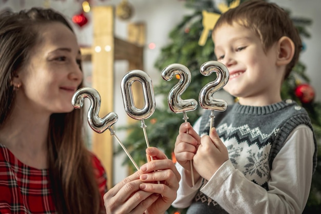 Feliz mamá e hijo en el fondo de un árbol de Navidad sostienen en sus manos los números plateados 2022 en palos. Concepto del año nuevo, ambiente festivo, decoración del hogar, felicitaciones.