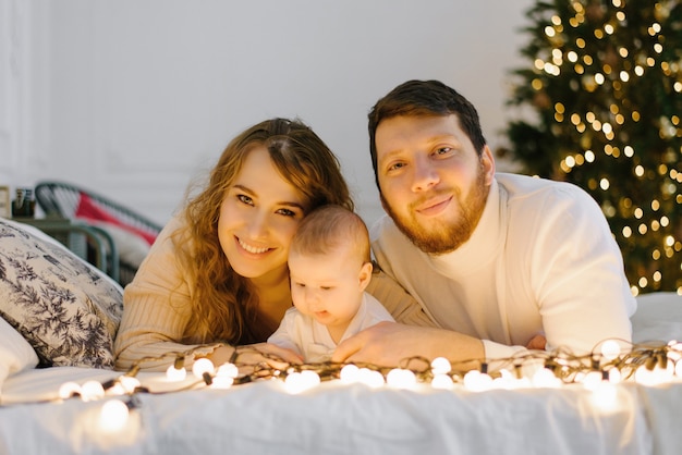 Feliz mamá y papá se acuestan en la cama junto a su bebé en el fondo de las luces de Navidad
