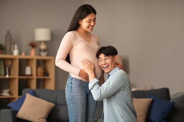 Feliz marido asiático ouvindo a barriga da esposa grávida em casa