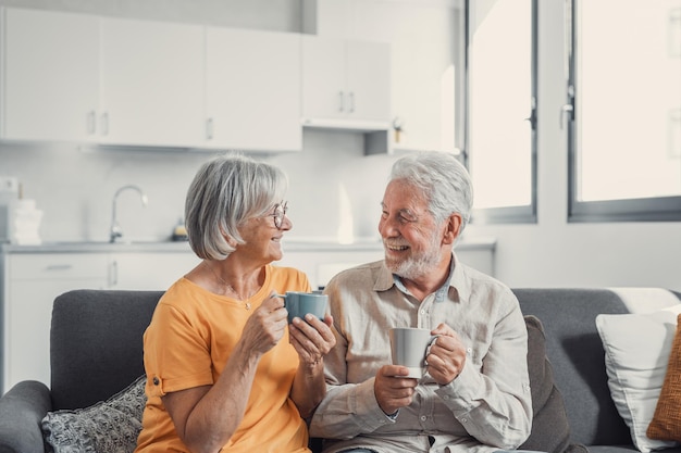 Feliz marido y mujer maduros de los años 50 se sientan a descansar en un cómodo sofá en la sala de estar disfrutan del té hablando sonriente pareja de ancianos de los años 60 relajarse en el sofá en casa beber café charlar hablar reír en el fin de semana de ocioxA