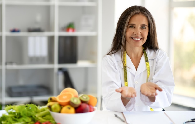 Feliz médica nutricionista europeia de meia-idade mantém espaço livre na mesa com frutas legumes