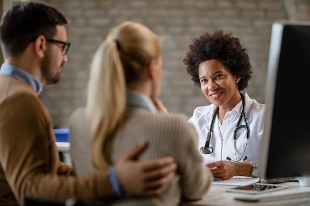 Feliz médico afro-americano conversando com um casal durante aconselhamento médico na clínica