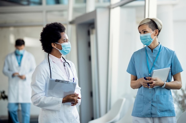 Feliz médico afro-americano e enfermeira falando enquanto usava máscaras protetoras em um corredor