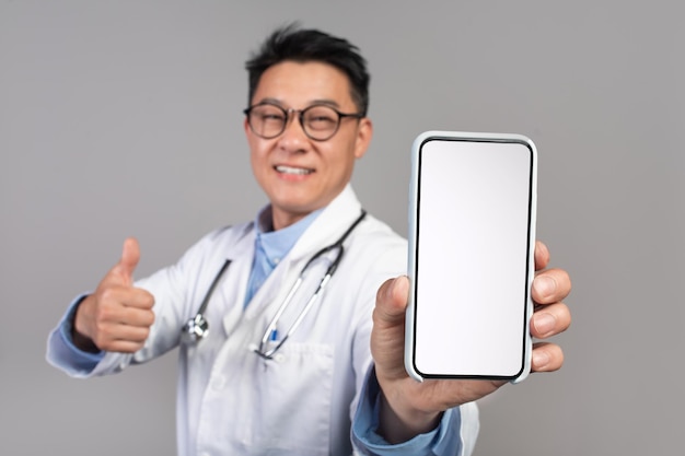 Feliz médico asiático maduro e confiante em óculos de jaleco branco mostra o telefone com o polegar da tela vazia para cima