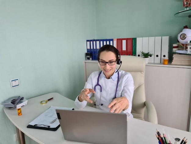 Feliz médico general atento en auriculares con computadora portátil y brindando ayuda en línea