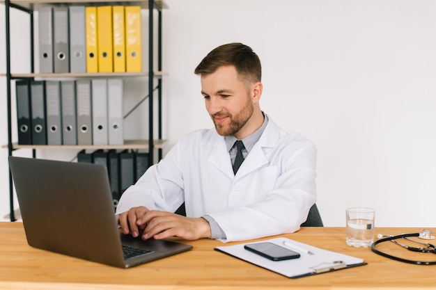 Feliz médico masculino viendo un seminario web médico en línea o un seminario de capacitación mientras se sienta con una computadora portátil en el lugar de trabajo