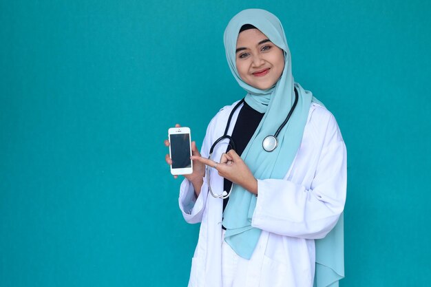 Feliz médico muçulmano asiático ou enfermeira com estetoscópio sorrindo e mostrando smartphone sobre fundo verde