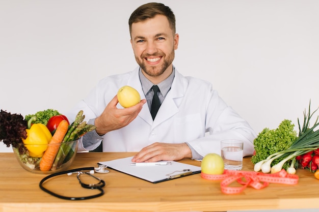 Foto feliz médico nutricionista sentado à mesa do local de trabalho entre legumes frescos e segurando a maçã