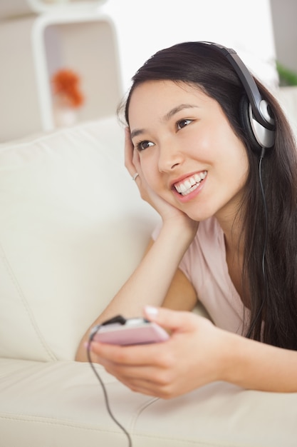 Feliz menina asiática deitada no sofá e ouvindo música com smartphone