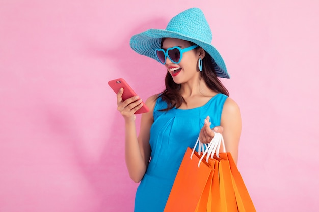 Feliz menina bonita asiática segurando sacolas de compras e telefone inteligente a desviar o olhar no fundo rosa