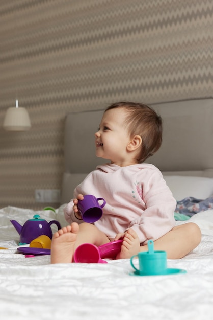 Foto feliz menina bonita brincando e fingindo beber chá de copos de brinquedo na cama