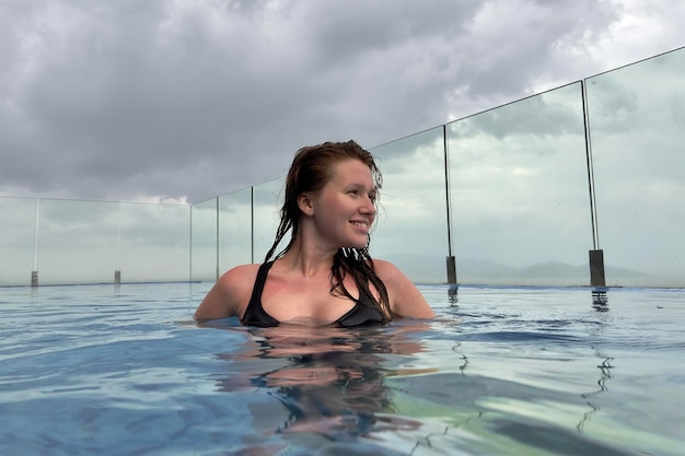 Feliz menina bonita e positiva jovem está aproveitando as férias de verão na piscina spa de luxo no telhado do hotel em um país tropical exótico com vista para o mar oceânico respira profundamente o ar fresco