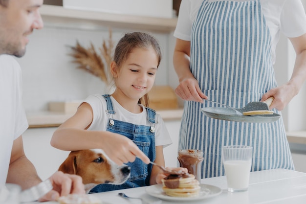 Feliz menina pequena gosta de comer sobremesa saborosa preparada pela mãe adiciona chocolate derretido a panquecas gosta de estar juntos e mãe pai e cachorro têm um delicioso café da manhã nutritivo na cozinha